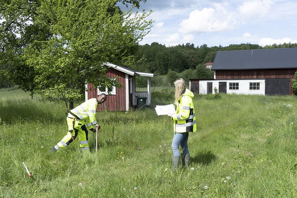 Två lantmätare utför en mätning på en grönskande tomt. I bakgrunden syns två stugor.