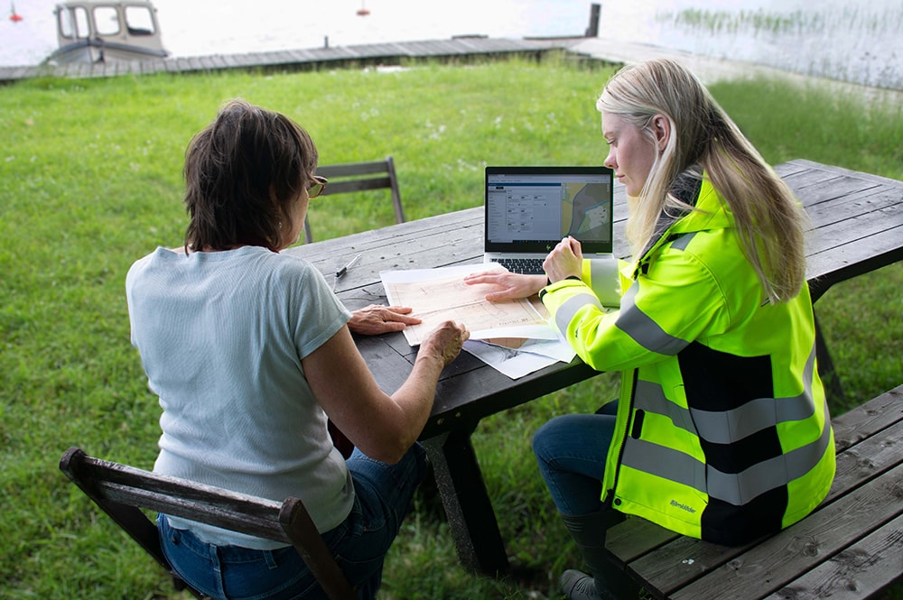 En lantmätare och en kund tittar på ett papper. De sitter utomhus vid ett bord. På bordet står en dator.